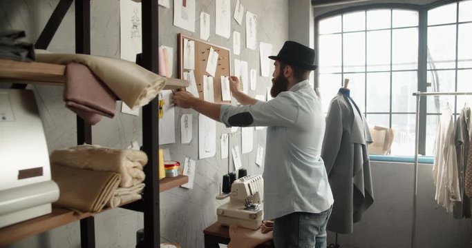 A male hipster clothes designers works with his sketches, looking at them pinned to wall, looking for inspiration for his new collection - fashion concept 4k footage