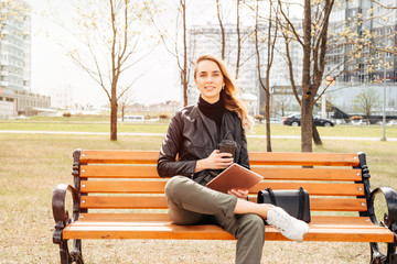 Young blonde woman sitting on park bench using tablet computer and drinking coffee