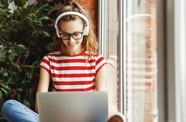 Happy young woman in headphones listening to music and working on laptop at home