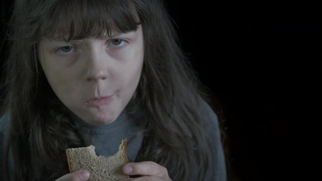 Homeless child with bread. Dirty sad little girl eats bread in the dark. Starving children concept.