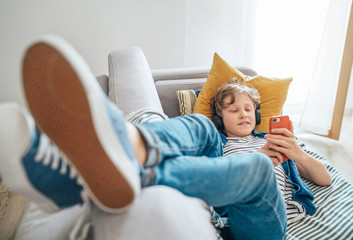 Preteen boy lying at home on cozy sofa dressed casual jeans and new sneakers listening to music and...