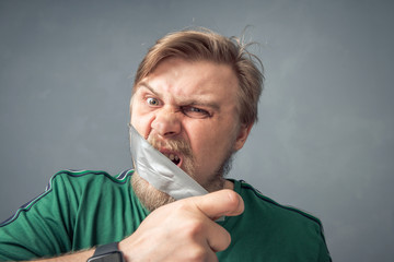 Close-up portrait of a bearded man ripping the tape off his face. The concept of what can't be said...