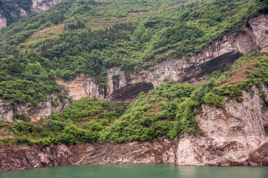 Zigui, China - May 6, 2010: Xiling gorge on Yangtze River. Large caves in brown rock cliffs with green foliage on top above green water.