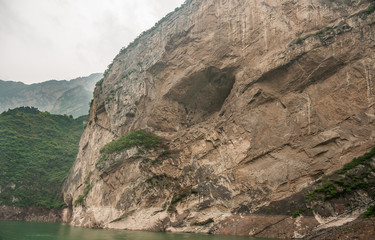 Xiangxicun, China - May 6, 2010: Xiling gorge on Yangtze River. Large cave hole in brown rock cliff descending in green water. Green covered mountains as backdrop. Patch of silver sky.