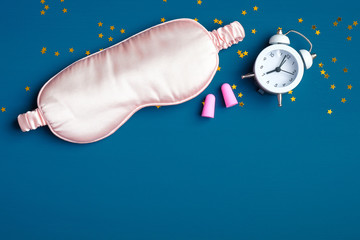 Healthy sleeping concept. Sleep eye mask with pink ear plugs and alarm clock on dark blue...