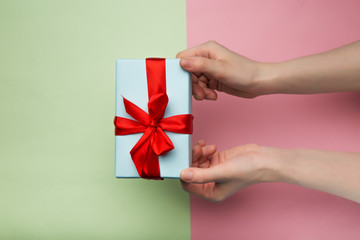 Top view of a woman's hands holding a white gift box with a red ribbon on a pink and green background. Flat lay. Gift for birthday, Christmas, Valentine's day, New year. Copy space