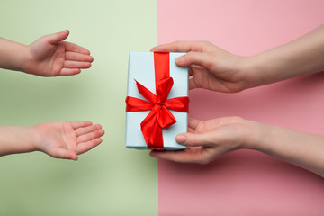 Top view of children's and women's hands holding a white gift box with a red ribbon on a pink and green background. Flat lay. Gift for birthday, Christmas, Valentine's day, New year. copy space.