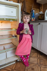 Hungry little girl in old clothes standing next to an empty refrigerator