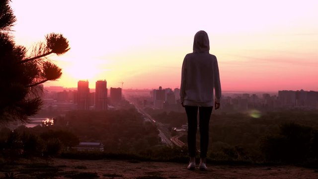 silhouette woman in casual sneakers and hoodie looking on the cityscape at sunrise rising hands urban city view in the morning