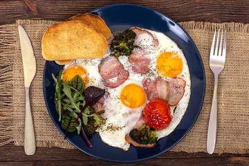 Breakfast or lunch with fried eggs, bread toast, arugula leaves, tomato, pork ham or bacon and broccoli on a dark blue plate with fork and knife, top view