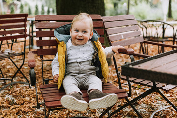 1 year old cute boy in stylish yellow jacket smiling and sitting on the chair in the park