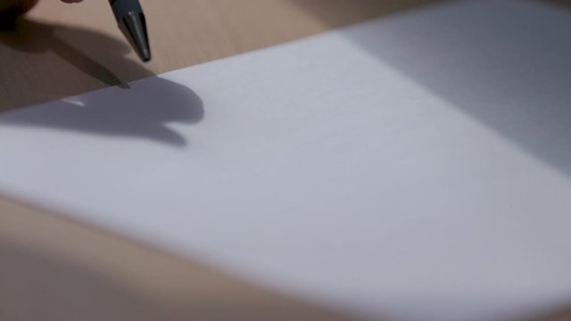 Man twirls and taps pen on blank piece of paper, shadow of hand and pen fall over paper. Writer's block, procrastination.