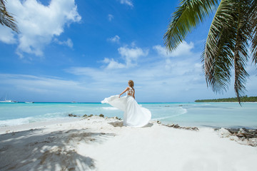 Fototapeta na wymiar Bride in a white wedding dress with a veil walking on the sandy caribbean beach at sunny day in Dominican republic 