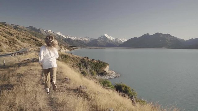 Blonde girl running beside a blue lake with snowy mountains in the background during sunrise in slow motion.