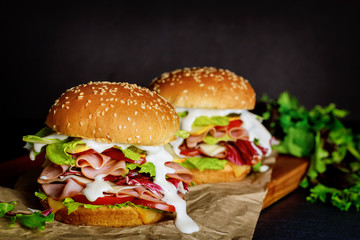 Two sandwiches with ham, cheese, tomatoes, lettuce on dark background.