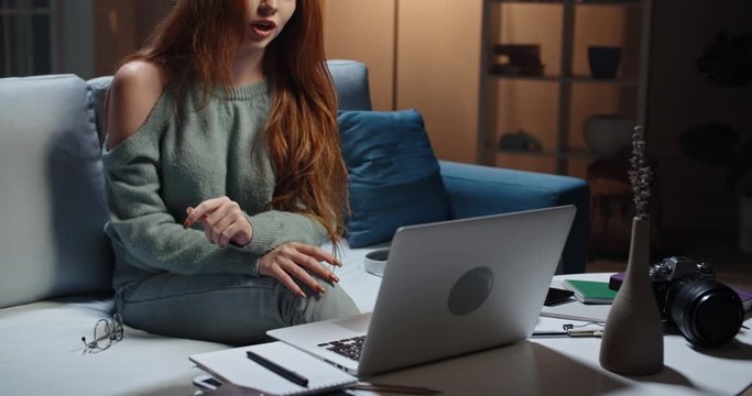 Pretty caucasian redhead girl sitting on coach looking at laptop, having a videochat or online tutorial, recording an internet vlog, cute model looking at webcamera and talking 4k footage