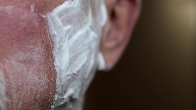 Senior man with essential tremor in hand shaves whiskers on his cheek with a safety razor, in close up slow motion clip.