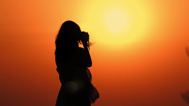 back view silhouette woman holding camera taking photos at sunrise warm sky with rising sun
