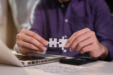 Business partnership concept. Businessman hand connecting jigsaw puzzle with calculator at office in morning light.