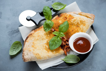 Calzone pizza with prosciutto, mozzarella and green basil over grey concrete background, studio shot