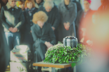 A metal urn with ashes of a dead person on a funeral, with people mourning in the background on a memorial service. Sad grieving moment at the end of a life. Last farewell to a person in an urn. - obrazy, fototapety, plakaty