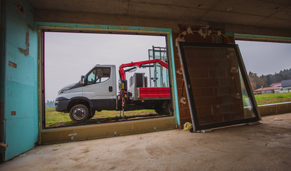 A truck with hydraulic arm and loaded with glass windows is waiting outside of a building and ready for lifting windows into the house. View from inside the house.
