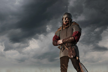 Warrior Viking in full arms with axe helmet and shield on dark sky background. Medieval scandinavian berserk red beard and fur costume attacks enemy. Concept historical photo