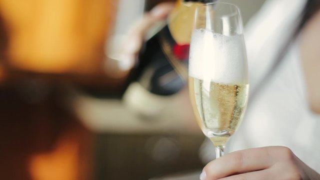Close-up shot of champagne pouring into glass in the hands of a young caucasian bride in a wedding dress. European girl pours herself a glass of champagne to quench her thirst, focusing on the glass.