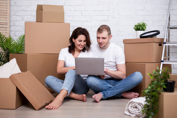 happy couple looking at laptop sitting in new house surrounded with cardboard boxes