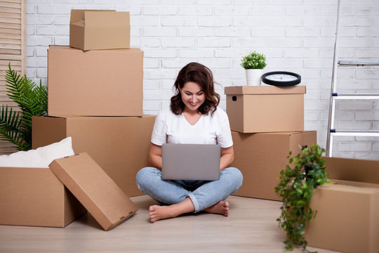 moving day - young woman using laptop in new house or flat surrounded with cardboard boxes