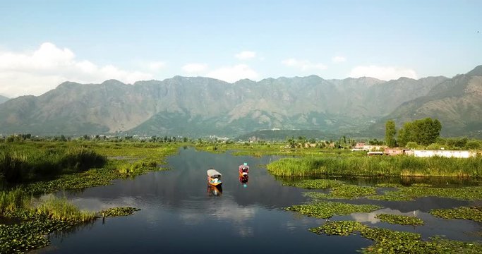 Cinematic aerial fly over of two traditional Shikaras floating on waterway at silent Dal Lake with panoramic mountainious landscape of Kashmir, India in the background.