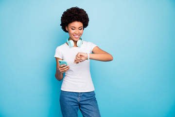 Photo of pretty dark skin curly lady hipster hold telephone app hands cool modern earphones check time watches meet friends wear casual white t-shirt jeans isolated blue color background