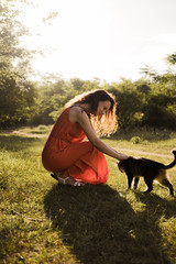 Beautiful young girl with dark curly hair in a bright orange dress strokes a fluffy spotted cat in the autumn forest.