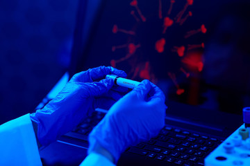 medicine, healthcare and virus concept - close up of hand in protective medical glove holding beaker with coronavirus positive blood test over laptop computer screen at laboratory