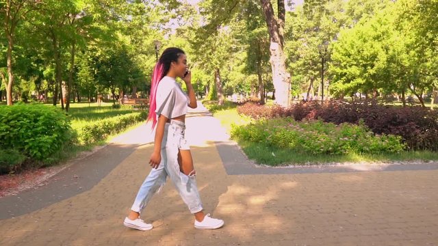 side view young african american woman talking by cell phone walking on the street in city park slow motion green landscape outdoors