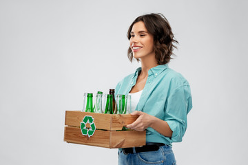 recycling, waste sorting and sustainability concept - smiling young woman holding wooden box with glass bottles and jars over grey background