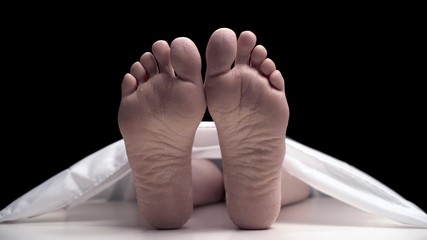 The naked corpse in the morgue lies on the table under a white sheet and moves its fingers. The skin on the legs is very dry and lifeless.
