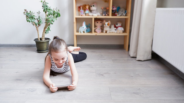 Distance learning. Little girl lying on a wooden floor with a phone, watching a cartoon or making a video call on a computer