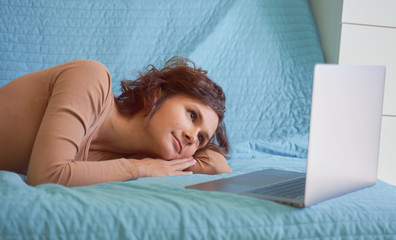 Happy young brunette girl is resting on comfortable sofa, using laptop at home, communicating with friends on social network, spending lazy weekends, watching movies TV series shopping online, emails