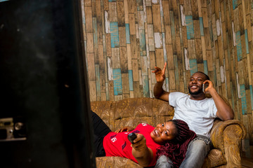 two young African couple watching television celebrating in their living room . Male using mobile phone