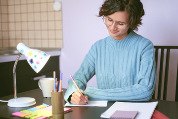 Portrait of young woman looking at letter friend, copywriter writes text in cozy home environment, smiling tenderly. Beautiful brunette girl with Bob hairstyle wears blue sweater. On table coffee mug