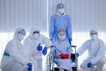 Old woman patient hold mini heart on a wheelchair with background of the medical team thumbs up after recovered from coronavirus or COVID19. 
Concept of success discover vaccine and teamwork.