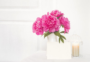 Pink peonies in white enamelled vase. Beautiful flowers in interior design. White paper for invitation text, white peonies in a vase, small bird case with candle in a middle, interior decoration.
