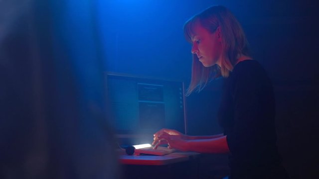 Attrctive woman hacker programmer is working on a computer in a dark underground office room. Blue neon light fills the room. She enters the software to steal login information.