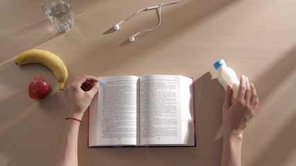 Woman is reading a book at the table, top view. On the table is a book, fruits and yogurt. Stay...