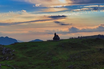 Fototapeta premium Little church in Velika Planina. Christian church in Big Pasture Plateau with sunrise colorful sky and beautiful mountain silhouettes in the background.