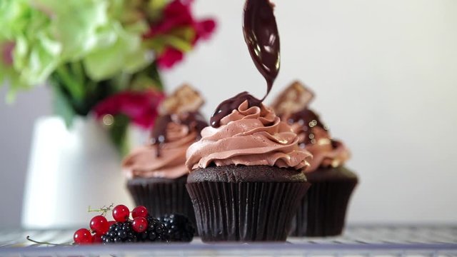 The process of putting chocolate coating on the top of the cupcake using spoon. Cooking delicious dessert, pastry. Chocolate brown sweet cake. Summer berries are laying next to cakes on the plate. 
