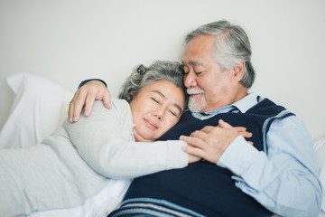 Senior Asian couple love lying on the bed with hug and hold the hand. Retired man and woman sleeping with talking on bed in bed room at home which smiling and felling happy. Old couple love concept