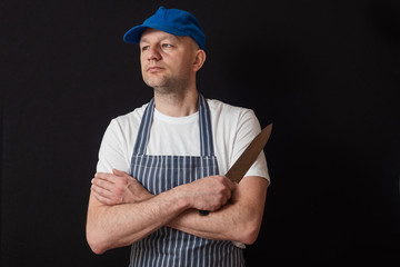 Portrait of a professional butcher in black and white apron, white t shirt and blue baseball hat, armed crossed, knife in his right hand. Meat industry concept.