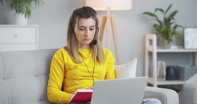 Female student processing a new lesson topic with an online tutor, listening to the teacher through headphones and writing information in a beautiful red notebook.